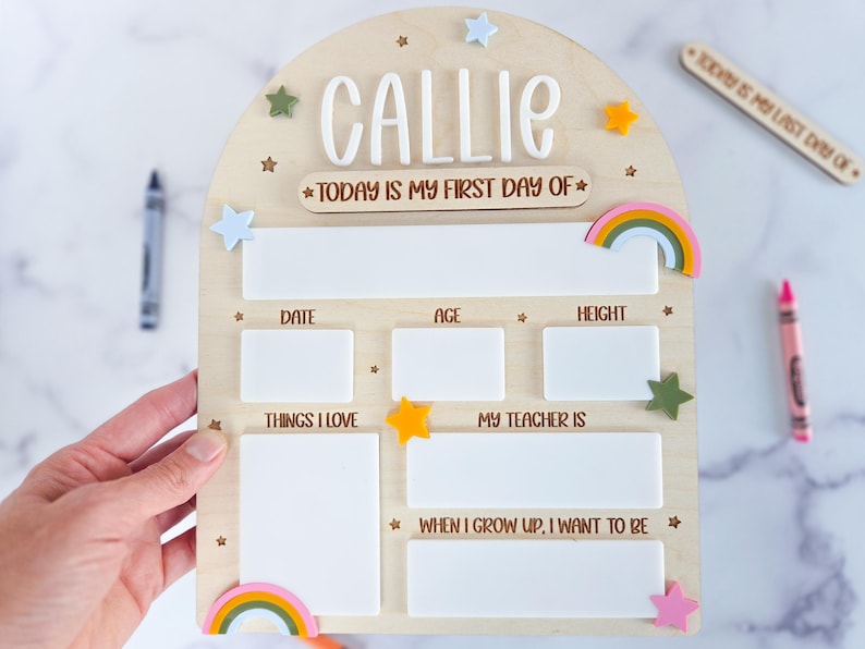 May include: An arched wooden sign with the name CALLIE in white 3D letters and the text TODAY IS MY FIRST DAY OF. It features white rectangular spaces for writing details like date, age, height, things I love, teacher, and future goals, decorated with small colorful stars and rainbows.