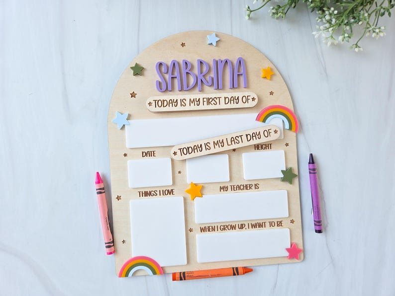 May include: Wooden school board with the name "Sabrina" in purple letters. Includes spaces for date, height, and other details. Decorated with stars, rainbows, and crayons. A pink and purple crayon are on either side.