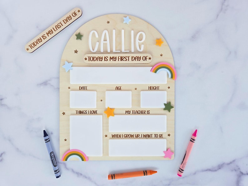 Personalized wooden back-to-school milestone board with the name "Callie" at the top. The board features interchangeable headers for "Today is my first day of" and "Today is my last day of," along with labeled sections for writing the date, age, height, teacher's name, favorite things, and future aspirations. Decorated with colorful stars and rainbows, the board includes white acrylic sections for dry-erase use. Surrounded by crayons on a white marble background.