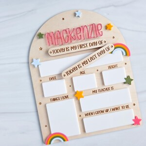 May include: Wooden first and last day of school board with the name "Mackenzie" in pink. Includes spaces for date, age, height, things I love, teacher, and future aspirations. Decorated with stars and rainbows.