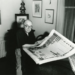 May include: A black and white photo of a man sitting in a chair, holding a large, unfolded document. The document is printed with text and illustrations. The man is wearing glasses and a dark shirt. The photo is taken in a room with a radiator and framed artwork on the walls.