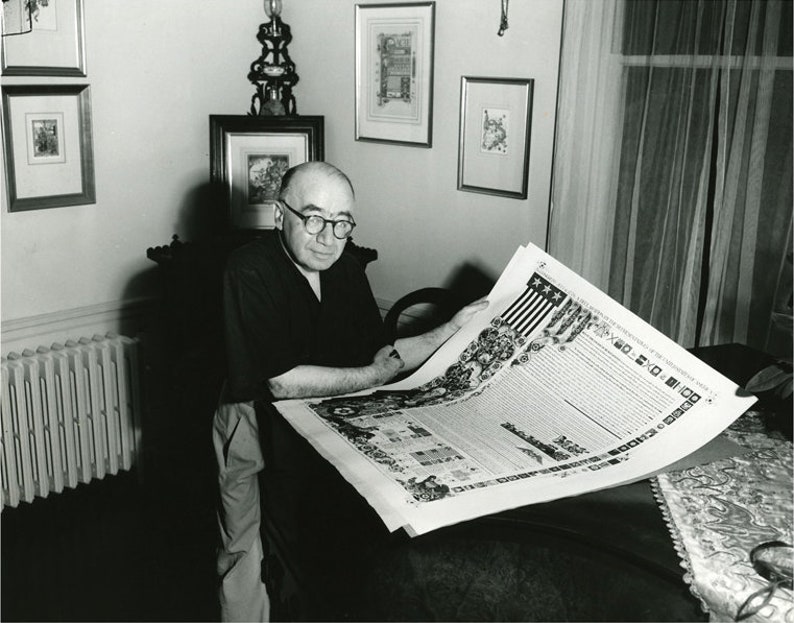 May include: A black and white photo of a man sitting in a chair, holding a large, unfolded document. The document is printed with text and illustrations. The man is wearing glasses and a dark shirt. The photo is taken in a room with a radiator and framed artwork on the walls.