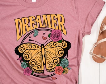 Könnte beinhalten: Ein mauvefarbenes T-Shirt mit einem Aufdruck eines gelben Schmetterlings mit bunten Blumen und dem Wort "DREAMER" in einer Retro-Schrift.
