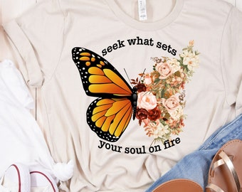 Könnte beinhalten: Ein beiges T-Shirt mit einem Aufdruck eines Schmetterlings und Blumen. Der Text "seek what sets your soul on fire" ist auf das T-Shirt gedruckt.