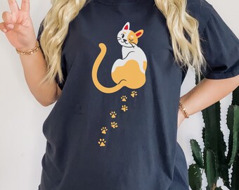 May include: Dark navy blue t-shirt featuring a cartoon cat with white and orange fur, winking and sitting. The cat has a long yellow tail and is followed by a trail of yellow paw prints. The shirt is a casual style.