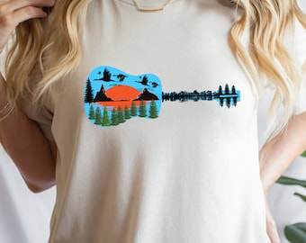 Op de afbeelding: Een wit T-shirt met een afbeelding van een gitaarsilhouet. De gitaar is gevuld met een landschapsscène met bergen, een meer, bomen en vogels die over een ondergaande zon vliegen.