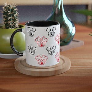 Może przedstawiać: Biały ceramiczny kubek z czarnym wnętrzem i uchwytem, ozdobiony czarnymi i czerwonymi twarzami Myszki Miki i Myszki Minnie. Kubek stoi na drewnianej podstawce na drewnianym stole. Kaktus w zielonej doniczce znajduje się w tle.