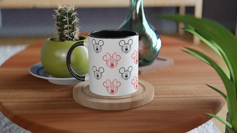 Może przedstawiać: Biały ceramiczny kubek z czarnym wnętrzem i uchwytem, ozdobiony czarnymi i czerwonymi twarzami Myszki Miki i Myszki Minnie. Kubek stoi na drewnianej podstawce na drewnianym stole. Kaktus w zielonej doniczce znajduje się w tle.