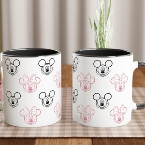 K&ouml;nnte beinhalten: Zwei wei&szlig;e Keramikbecher mit schwarzen R&auml;ndern und silbernen Griffen. Die Becher sind mit einem Muster aus Mickey- und Minnie-Mouse-Gesichtern in Schwarz und Rosa verziert. Die Becher stehen auf einer karierten Oberfl&auml;che.