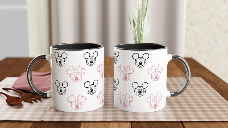 K&ouml;nnte beinhalten: Zwei wei&szlig;e Keramikbecher mit schwarzen R&auml;ndern und silbernen Griffen. Die Becher sind mit einem Muster aus Mickey- und Minnie-Mouse-Gesichtern in Schwarz und Rosa verziert. Die Becher stehen auf einer karierten Oberfl&auml;che.