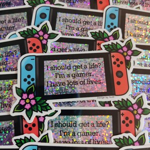 May include: A collection of stickers featuring a stylized video game console design. The stickers have a black background with a holographic effect. Each sticker has a console with a blue and red controller, floral accents, and the text: "I should get a life? I'm a gamer. I have lots of lives."