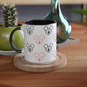 K&ouml;nnte beinhalten: Eine wei&szlig;e Keramik-Tasse mit schwarzem Innenraum und Henkel, verziert mit schwarzen und rosa Cartoon-Mausgesichtern. Die Tasse steht auf einem Holzuntersetzer auf einem runden Holztisch. Ein kleiner Kaktus in einem gr&uuml;nen Topf befindet sich im Hintergrund.