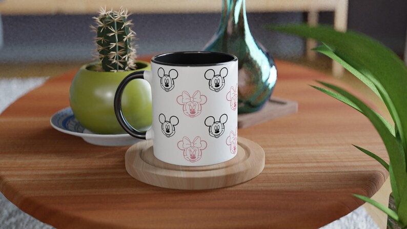 K&ouml;nnte beinhalten: Eine wei&szlig;e Keramik-Tasse mit schwarzem Innenraum und Henkel, verziert mit schwarzen und rosa Cartoon-Mausgesichtern. Die Tasse steht auf einem Holzuntersetzer auf einem runden Holztisch. Ein kleiner Kaktus in einem gr&uuml;nen Topf befindet sich im Hintergrund.