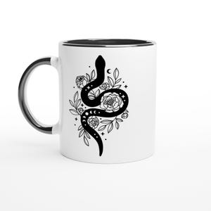 Peut inclure: Un mug en céramique blanche avec un intérieur et une anse noirs. L'extérieur présente une illustration graphique noire d'un serpent enroulé entouré d'éléments floraux, de feuilles, de petites étoiles et de symboles de phases lunaires.