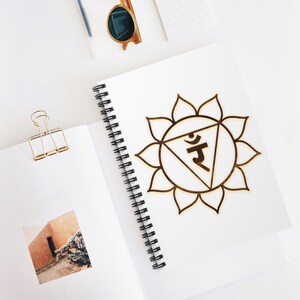 May include: A white spiral bound notebook with a gold mandala design on the cover. The mandala is a symbol of the solar plexus chakra, which is associated with personal power and self-esteem. The notebook is perfect for journaling, taking notes, or sketching.