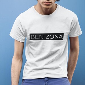 May include: White t-shirt with the text "BEN ZONA" in a black rectangle. The shirt has short sleeves and a crew neck. The background is a solid light blue.
