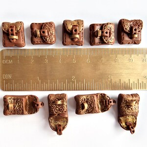 Op de afbeelding: Tien miniatuur bruine leren tassen met gouden accenten en gespen. De tassen zijn ongeveer 2,5 cm groot en zijn gerangschikt in twee rijen op een wit oppervlak. Een liniaal is zichtbaar op de achtergrond.