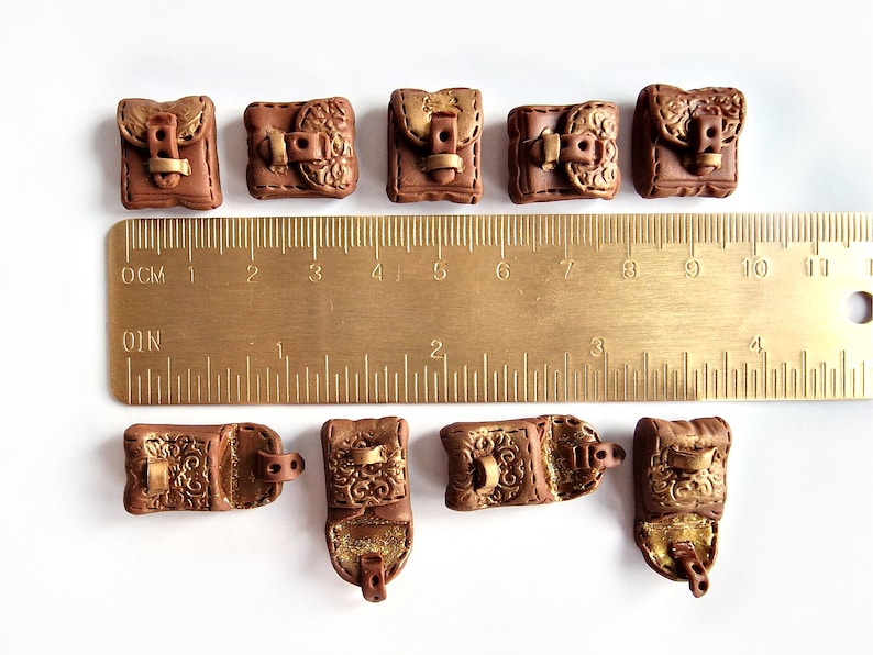 Op de afbeelding: Tien miniatuur bruine leren tassen met gouden accenten en gespen. De tassen zijn ongeveer 2,5 cm groot en zijn gerangschikt in twee rijen op een wit oppervlak. Een liniaal is zichtbaar op de achtergrond.