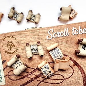 May include: A set of ten miniature scroll tokens, each with a different design and inscription. The scrolls are made of a light brown material and have a slightly textured surface. The tokens are arranged on a wooden surface with the words "Scroll tokens" written in white.