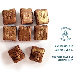 Op de afbeelding: Acht kleine, vierkante, bruine kleiboekcharms met gouden accenten en decoratieve details. De charms hebben een gestikte look rond de randen. De charms zijn handgemaakt en elk stuk is uniek. De tekst op de afbeelding luidt: "HANDGEMAAKTE ARTIKELEN ZIJN UNIEK * JE ZULT NOOIT TWEE IDENTIEKE STUKKEN VINDEN"