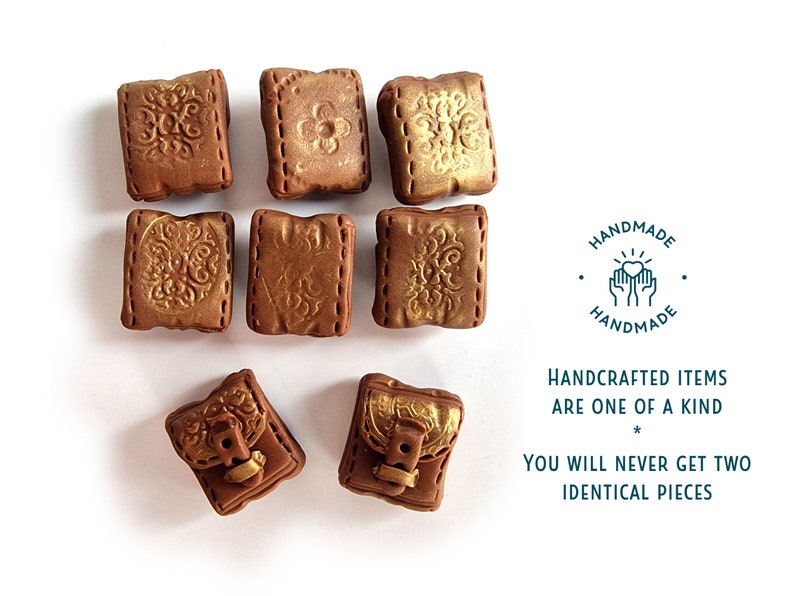 Op de afbeelding: Acht kleine, vierkante, bruine kleiboekcharms met gouden accenten en decoratieve details. De charms hebben een gestikte look rond de randen. De charms zijn handgemaakt en elk stuk is uniek. De tekst op de afbeelding luidt: "HANDGEMAAKTE ARTIKELEN ZIJN UNIEK * JE ZULT NOOIT TWEE IDENTIEKE STUKKEN VINDEN"