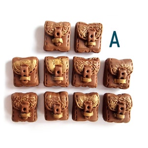Op de afbeelding: Negen bruine en gouden miniatuur tassen met gespen en stiksels. De tassen zijn in een piramidevorm gerangschikt. De letter "A" is zichtbaar in de rechterbovenhoek van de afbeelding.