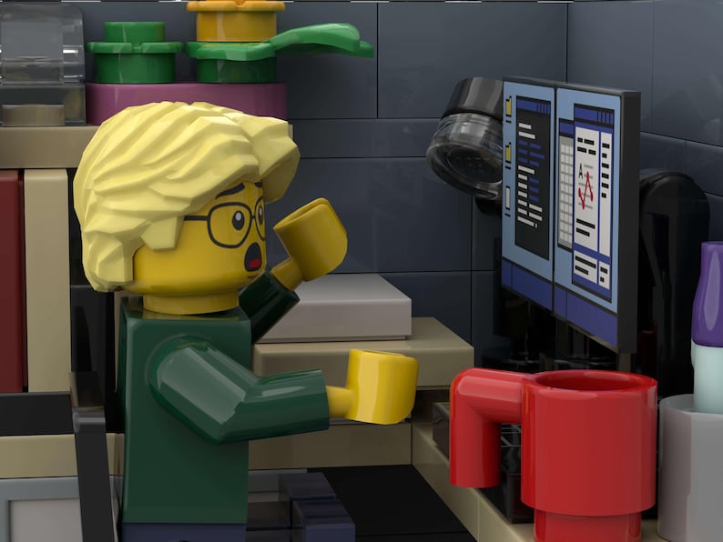 Puede incluir: Una figura de Lego con pelo rubio y gafas, sosteniendo dos tazas amarillas, parece sorprendida mientras mira una pantalla de ordenador. Una taza roja est&aacute; sobre un escritorio. La escena est&aacute; ambientada en un entorno de oficina Lego.