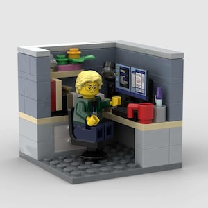 Może przedstawiać: Diorama Lego przedstawiająca biuro. Figurka Lego z blond włosami i okularami siedzi przy biurku z dwoma monitorami komputerowymi. Na biurku znajduje się czerwony kubek i klawiatura. Biuro jest szare z półką na książki i innymi akcesoriami.