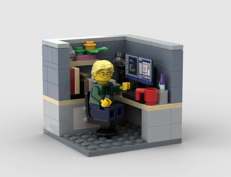 Puede incluir: Diorama Lego de un entorno de oficina. Una figura de Lego con pelo rubio y gafas est&aacute; sentada en un escritorio con dos monitores de ordenador. El escritorio tiene una taza roja y un teclado. La oficina tiene una estanter&iacute;a con libros y plantas.