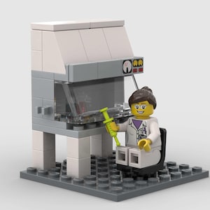 Puede incluir: Un laboratorio Lego blanco con una campana de plástico transparente y una figura de científica Lego femenina sosteniendo una jeringa. La científica lleva una bata de laboratorio blanca y tiene el pelo castaño y la cara amarilla.