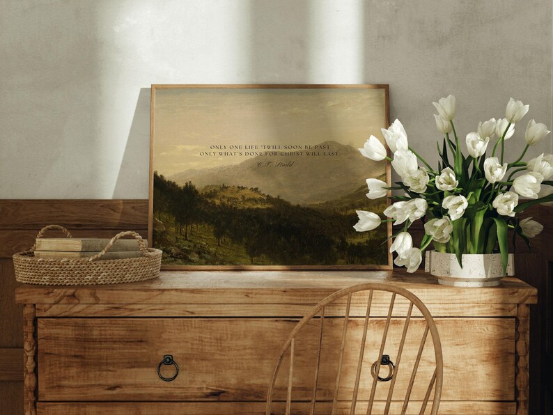 May include: A framed print with a quote by the author, "Only one life, twill soon be past, Only what's done for Christ will last." The print features a landscape painting of a mountain range with trees and a cloudy sky. The print is sitting on a wooden table with a vase of white tulips.
