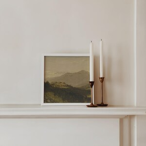 May include: A white framed print of a landscape painting with a quote in the center. The quote reads "Open your eyes and still you won't believe, only what a wilderness surrounds us all." - W.B. Yeats. Two white candles are on a gold candlestick holder on a white mantel.