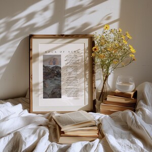 May include: A framed print with the text "ROCK OF AGES" and a landscape painting. A vase of yellow flowers, a glass of water, and a stack of books are on a white surface. The scene is bathed in sunlight.