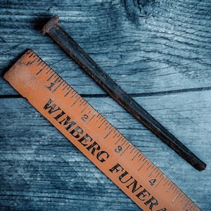 May include: A rusty, dark metal nail and an orange wooden ruler with black text. The ruler is marked with inch measurements and the text "WIMBERG FUNERAL HOME". The nail is positioned diagonally across the wooden surface.