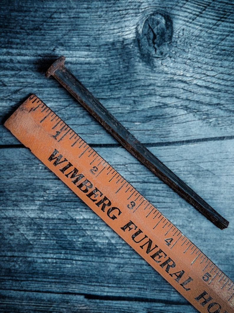 May include: A rusty, dark metal nail and an orange wooden ruler with black text. The ruler is marked with inch measurements and the text "WIMBERG FUNERAL HOME". The nail is positioned diagonally across the wooden surface.