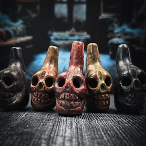 May include: Five ceramic skull figurines in a row on a dark wooden surface. The skulls are painted in different colors, including black, red, yellow, and brown. The skulls have a spooky and whimsical look.