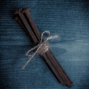 May include: A bundle of rusty, square-headed nails tied together with twine. The nails are a dark brown color and are arranged diagonally against a dark blue wooden background.