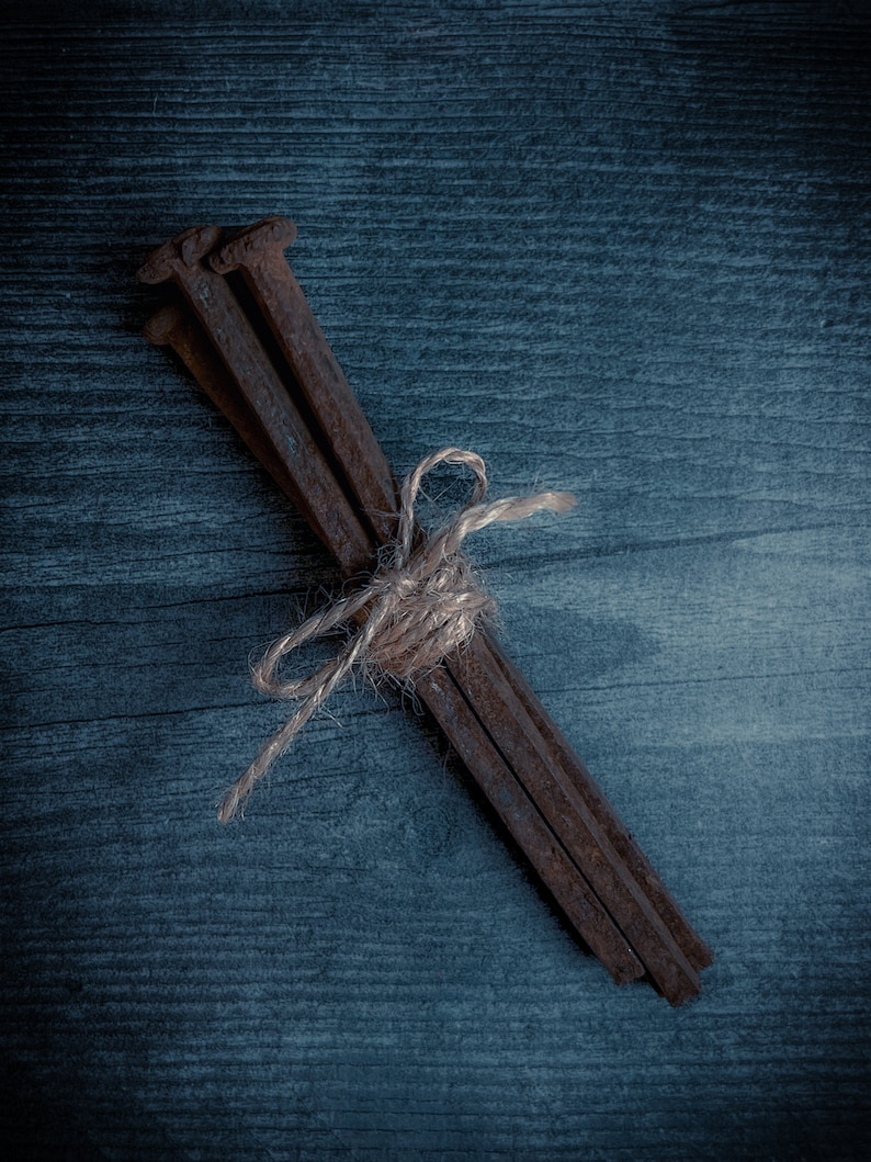 May include: A bundle of rusty, square-headed nails tied together with twine. The nails are a dark brown color and are arranged diagonally against a dark blue wooden background.