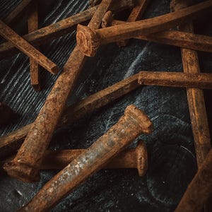 May include: Close-up of several rusty, square-headed nails scattered on a dark, textured surface. The nails are various sizes and show signs of age and oxidation, with a prominent reddish-brown color. The image has a vintage, industrial aesthetic.