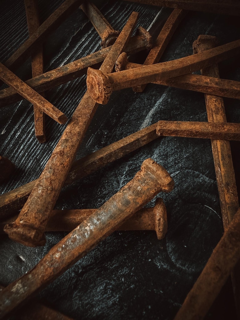 May include: Close-up of several rusty, square-headed nails scattered on a dark, textured surface. The nails are various sizes and show signs of age and oxidation, with a prominent reddish-brown color. The image has a vintage, industrial aesthetic.