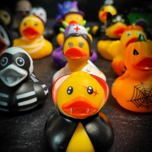 Könnte beinhalten: Eine Sammlung von Halloween-Gummienten. Die Enten sind als Vampir, Krankenschwester, Hexe, Skelett und Kürbis verkleidet. Die Enten sind gelb, schwarz, orange und weiß, mit bemalten Details. Die Vampir-Ente hat einen schwarzen Umhang und einen roten Kragen.