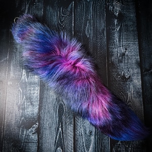 Puede incluir: Una cola de piel sintética en tonos azules, morados y rosas, con efecto degradado. La cola es esponjosa y tiene una ligera curva. Se ve un pequeño cierre dorado en la parte superior.