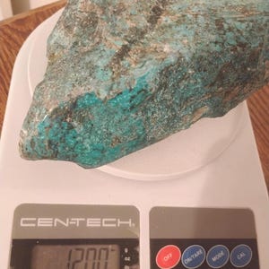 May include: A large, irregularly shaped turquoise-colored rock with dark brown and black inclusions, resting on a white digital scale. The scale displays a weight of 12.00 lbs. The scale is a Centech model.
