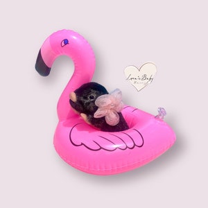 May include: A small black and white stuffed animal wearing a pink flower sits in a pink inflatable flamingo pool float. The float has a black beak and black eyes.