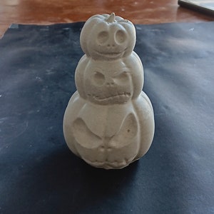May include: A white ceramic pumpkin stack with three pumpkins, each with a different carved jack-o'-lantern face.