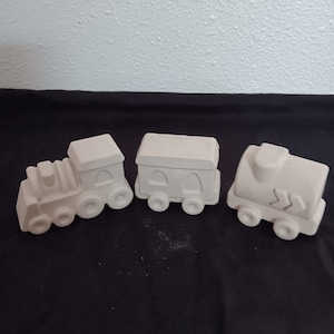 May include: Three ceramic train cars, painted white, ready to be decorated. The train engine has a large chimney and a rounded front. The middle car has two windows and the last car has a rounded back and a small door.