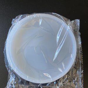 May include: A circular, translucent white silicone mold for crafting, featuring a smooth, flat interior surface and a raised outer rim. The mold is enclosed in a clear, crinkled plastic packaging sleeve that is sealed along the side edges.
