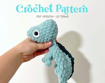 May include: A crocheted chameleon amigurumi toy in teal and dark green, with a white eye. The toy is being held by a hand.