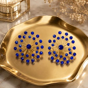 May include: A pair of circular earrings featuring a central deep blue bead surrounded by radiating gold-toned wire spokes, each tipped with a small, faceted royal blue bead, creating a sunburst design that resembles a firework or floral pattern.