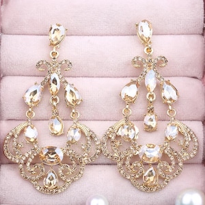 May include: A pair of gold-toned chandelier earrings with multiple clear and champagne-colored crystals. The earrings have an ornate design with a teardrop shape and are set against a pink background.